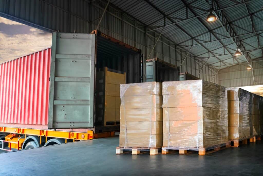 Palletized boxes wrapped in shrink wrap staged on a warehouse dock beside an open red shipping container trailer ready for loading.